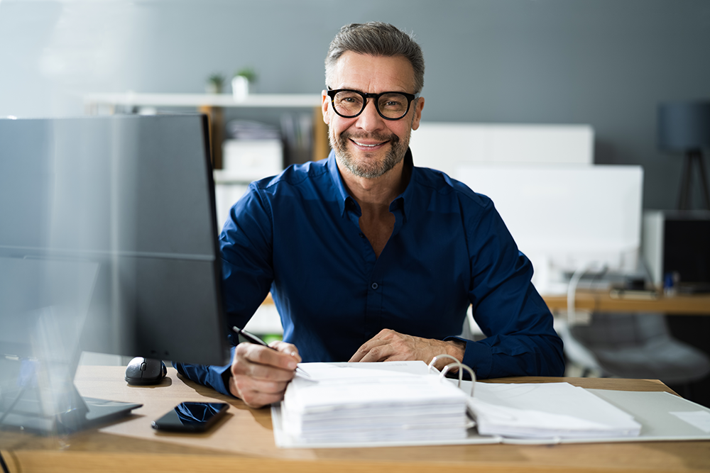 Professional man in front of computer smiling with stack of papers