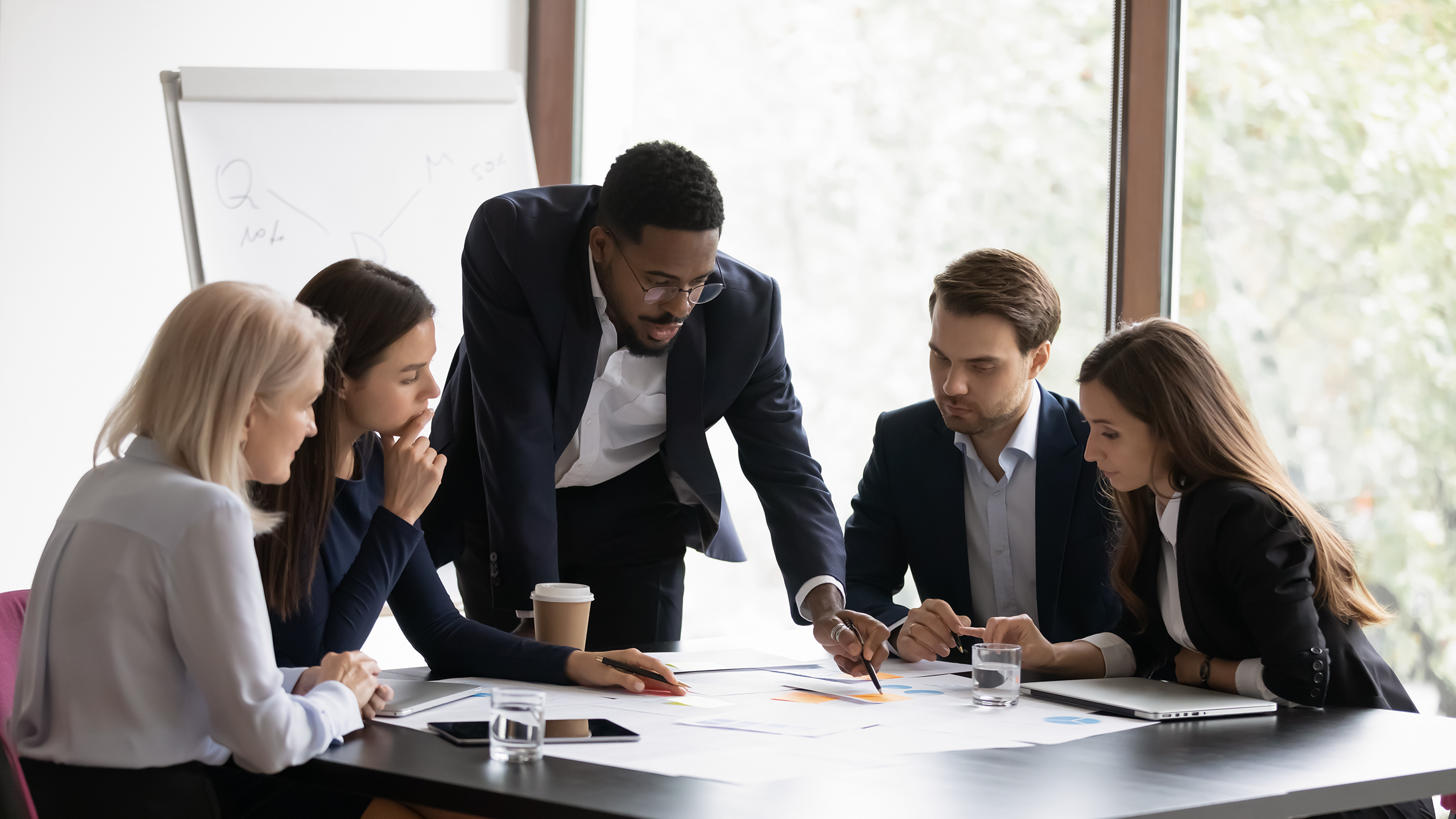 A professional coach stands at a table with four people, who are seated and who look at a piece of paper the coach is pointing at