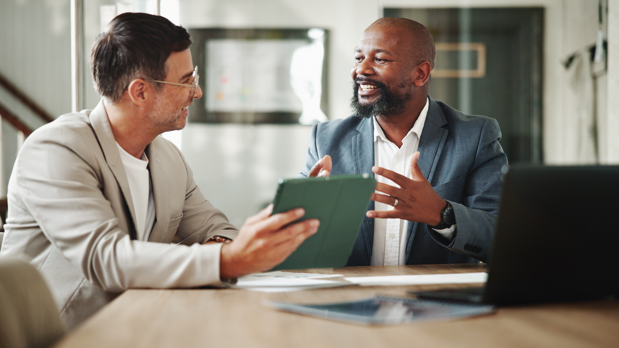 A professional coach sits at a table with his mentor, who holds up an iPad as the two men talk.