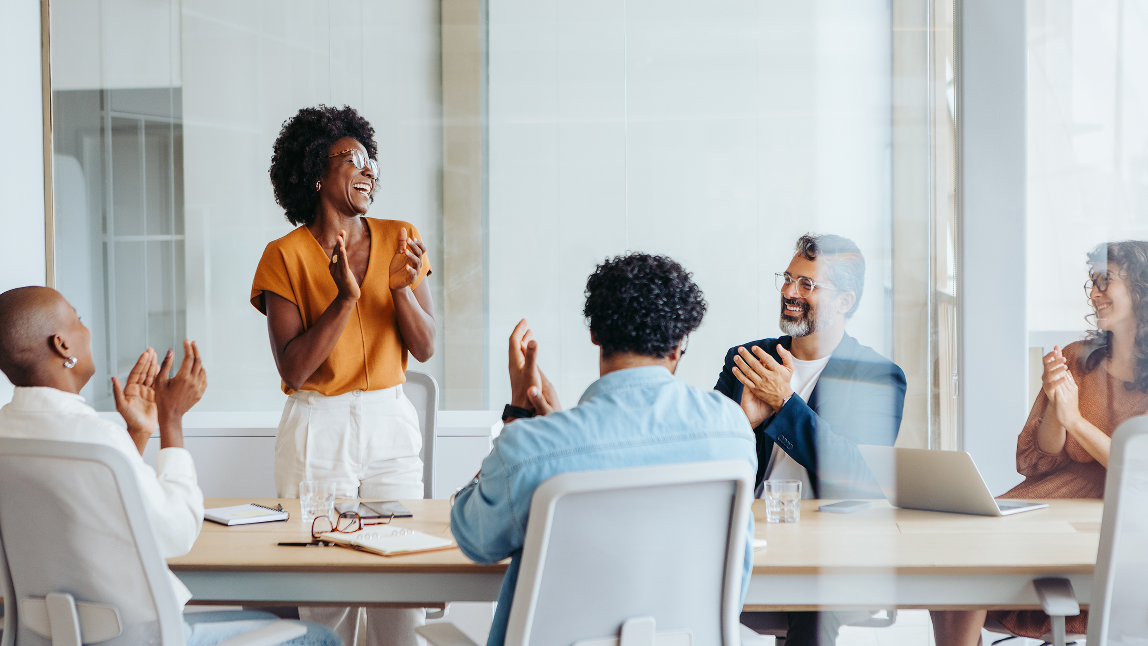 A professional female coach stands and addresses four employees who are sitting at a big table.