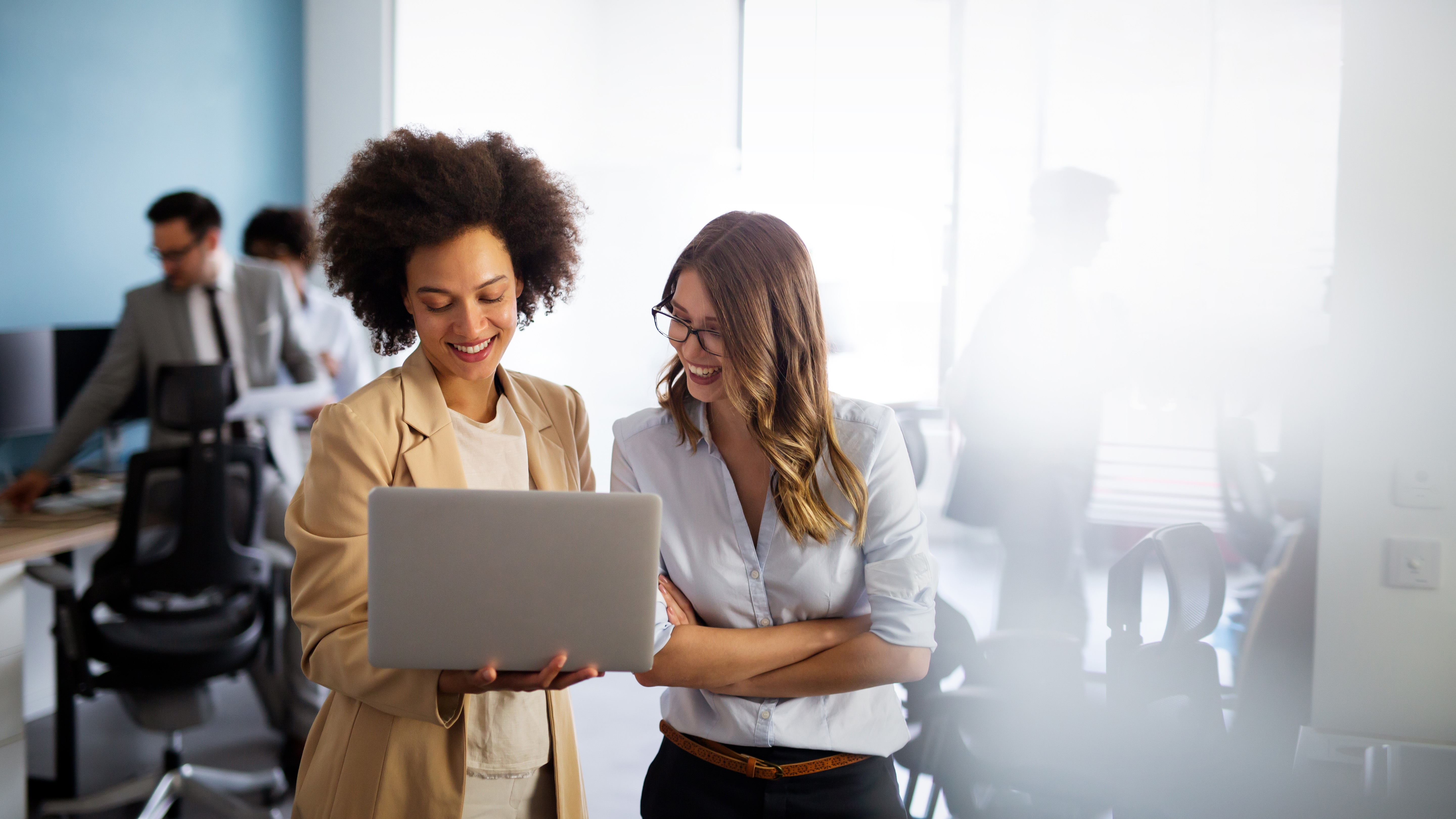 A coach stands with her mentee. The coach is holding an open laptop, which the mentee is looking at