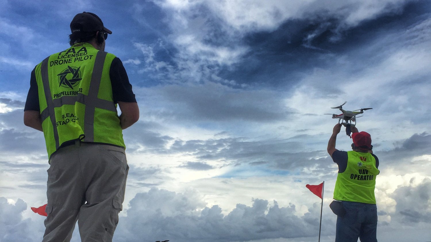 There are two drone pilots standing on the ground. They both wear headsets and yellow reflective vests. The pilot on the left watches the pilot on the right, who is launching a drone into the air.